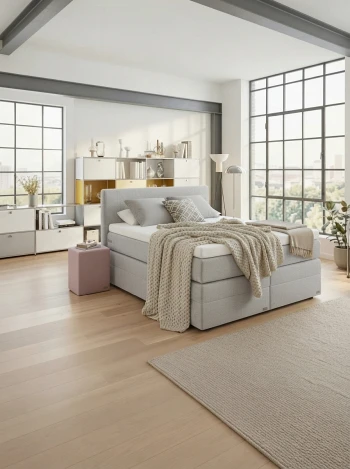 Hellgraues Boxspringbett in einem lichtdurchfluteten Loft-Schlafzimmer mit großen Industriefenstern und Zimmerpflanzen