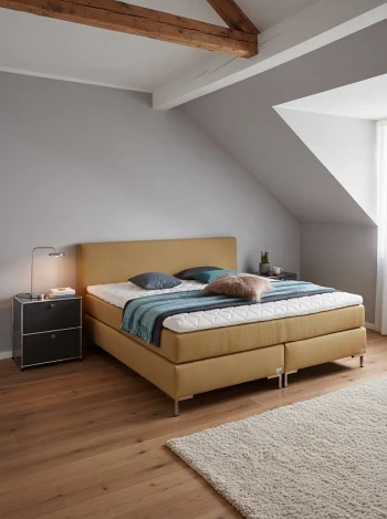 Senfgelbes Boxspringbett in einem gemütlichen Dachschlafzimmer mit rustikalen Holzbalken und gedämpftem Licht