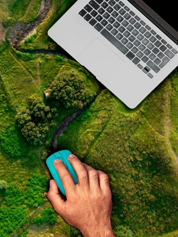 Hand mit türkisfarbener Computermaus neben einem Laptop auf einer Luftaufnahme mit Wiesen, Bäumen und einem Bachlauf