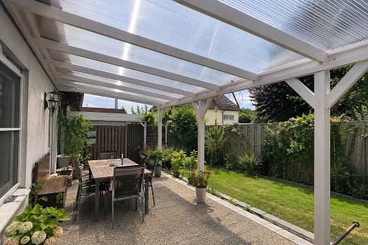Helle Terrassenüberdachung mit weißem Holzrahmen und transparenten Stegplatten, darunter Holz-Esstisch mit schwarzen Gartenstühlen und Gartenpflanzen