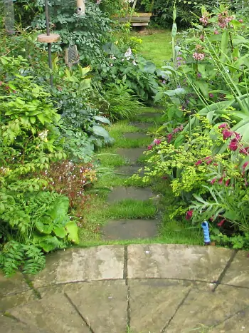 Gartenweg aus Naturstein-Trittplatten durch einen üppig bepflanzten Cottage-Garten mit Farnen, Hostas und roten Blüten, im Hintergrund eine Bank.